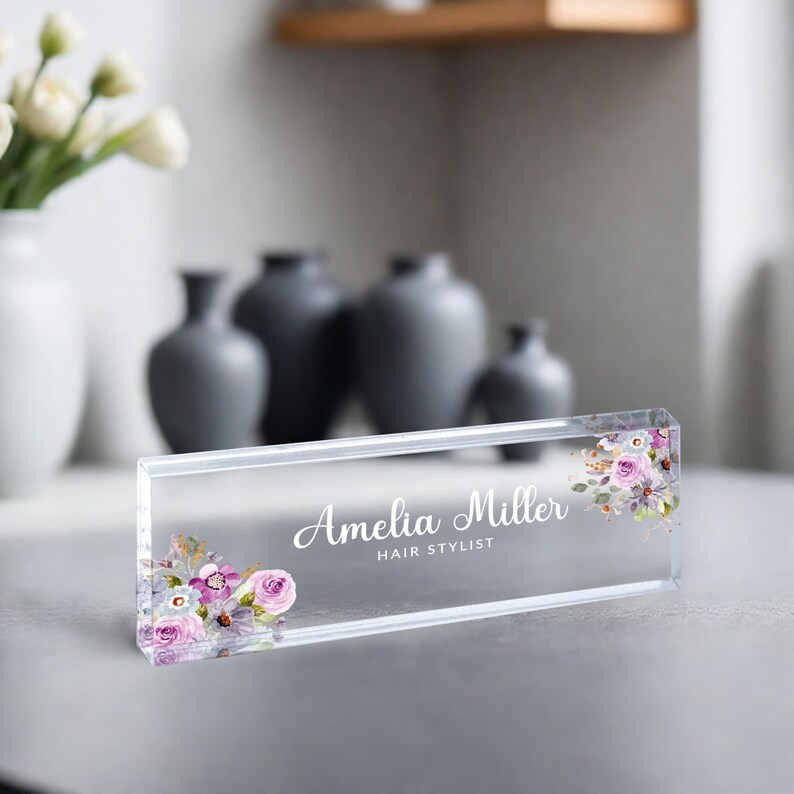 May include: Clear acrylic desk nameplate with the text "Amelia Miller Hair Stylist" and a floral design with pink and purple flowers.