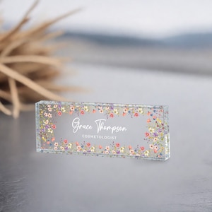 May include: A clear glass desk nameplate with a floral border and the text "Grace Thompson COSMETOLOGIST".