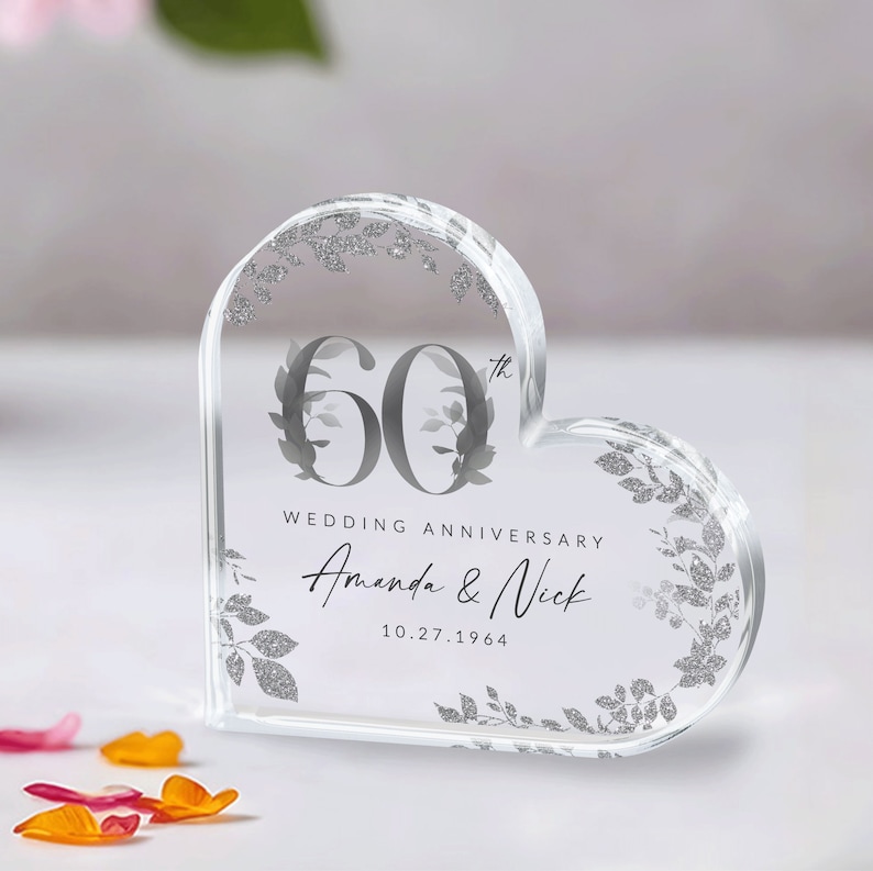 May include: A clear heart-shaped acrylic paperweight with a silver glitter design. The paperweight features the number 60 with a floral design and the text "Wedding Anniversary Amanda & Nick 10.27.1964".