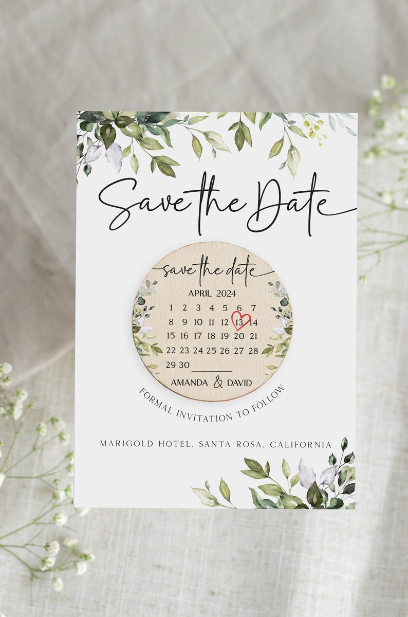 Puede incluir: Tarjeta de guardar la fecha con un calendario circular de madera que presenta un coraz&oacute;n rojo el 13 de abril. La tarjeta est&aacute; decorada con ilustraciones florales de acuarela verde y blanca. El texto dice "Save the Date" y "Amanda & David" con la ubicaci&oacute;n "Marigold Hotel, Santa Rosa, California".