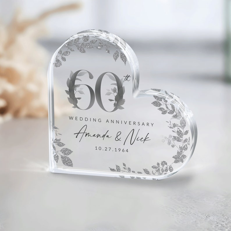 May include: A clear heart-shaped crystal paperweight with a silver glitter design. The paperweight features the number 60 with a 'th' superscript, the words 'Wedding Anniversary', and the names 'Amanda & Nick' with the date '10.27.1964'.