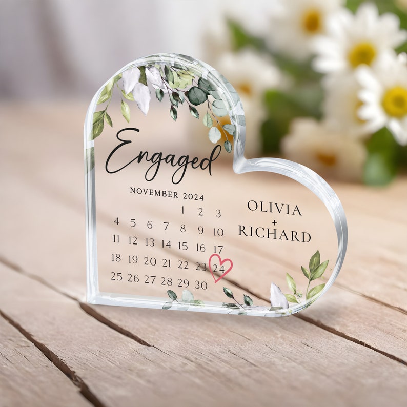 K&ouml;nnte beinhalten: Ein durchsichtiger, herzf&ouml;rmiger Acryl-Briefbeschwerer mit floralem Design und den Worten "Verlobt" und "Olivia + Richard". Der Briefbeschwerer enth&auml;lt auch einen Kalender f&uuml;r November 2024, wobei der 24. Tag umkreist ist und ein kleines rosa Herz.