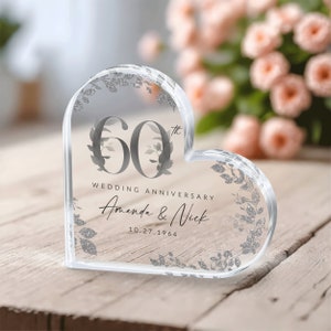 May include: A clear heart-shaped crystal paperweight with a silver glitter design. The paperweight features the text "60th Wedding Anniversary Amanda & Nick 10.27.1964".