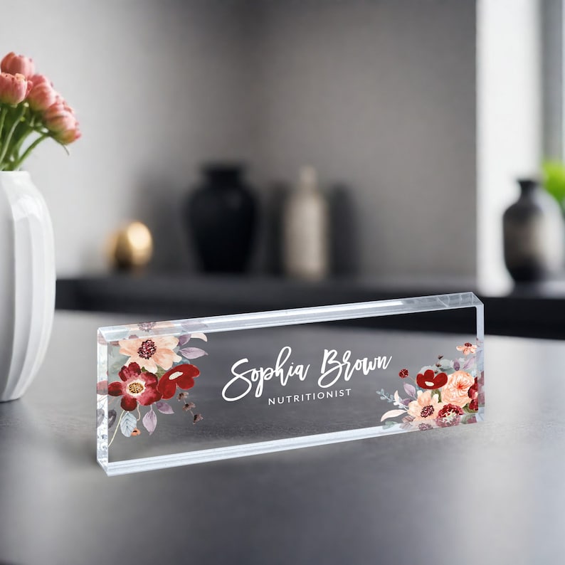 May include: A clear acrylic desk nameplate with a floral design in shades of red, pink, and white. The nameplate reads "Sophia Brown, Nutritionist".