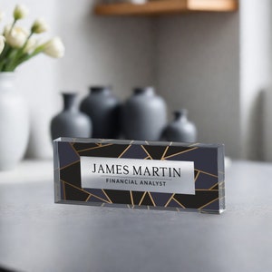 May include: A clear acrylic desk nameplate with a geometric pattern in gold and black. The nameplate reads "JAMES MARTIN FINANCIAL ANALYST".