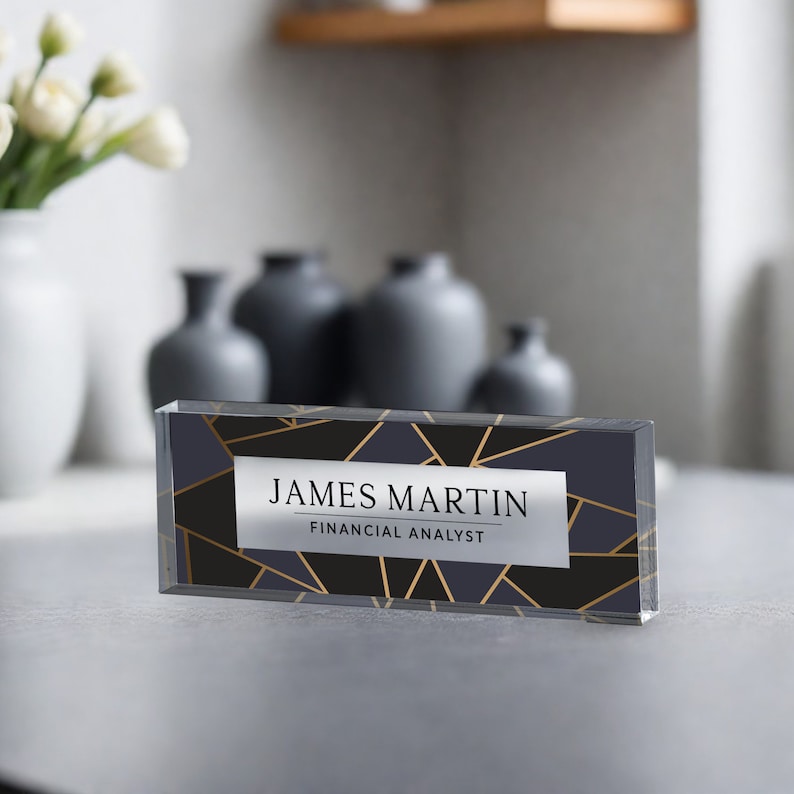 May include: A clear acrylic desk nameplate with a geometric pattern in gold and black. The nameplate reads "JAMES MARTIN FINANCIAL ANALYST".
