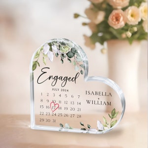 K&ouml;nnte beinhalten: Ein durchsichtiger, herzf&ouml;rmiger Kristall-Briefbeschwerer mit floralem Design und dem Text "Verlobt" und "Juli 2024". Der Kalender zeigt das Datum "17" in einem rosa Kreis. Die Namen "Isabella" und "William" sind ebenfalls enthalten.