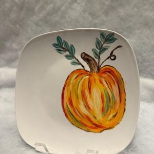 May include: A square white ceramic plate featuring a hand-painted pumpkin design. The pumpkin is orange, yellow, and red with green and brown accents. Green leaves and a brown stem complete the autumn-themed artwork.