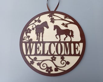 May include: A round wooden sign with a brown stain, featuring a welcome message and two horse silhouettes. The sign is decorated with a floral design and has a hanging loop.