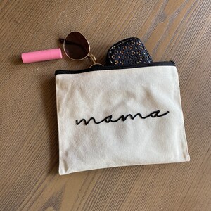 May include: A beige canvas zippered pouch with black embroidery that reads "mama". The pouch is lying on a wooden surface with a pair of sunglasses and a pink lipstick tube beside it.