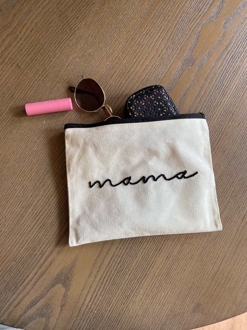 May include: A beige canvas zippered pouch with black embroidery that reads "mama". The pouch is lying on a wooden surface with a pair of sunglasses and a pink lipstick tube beside it.