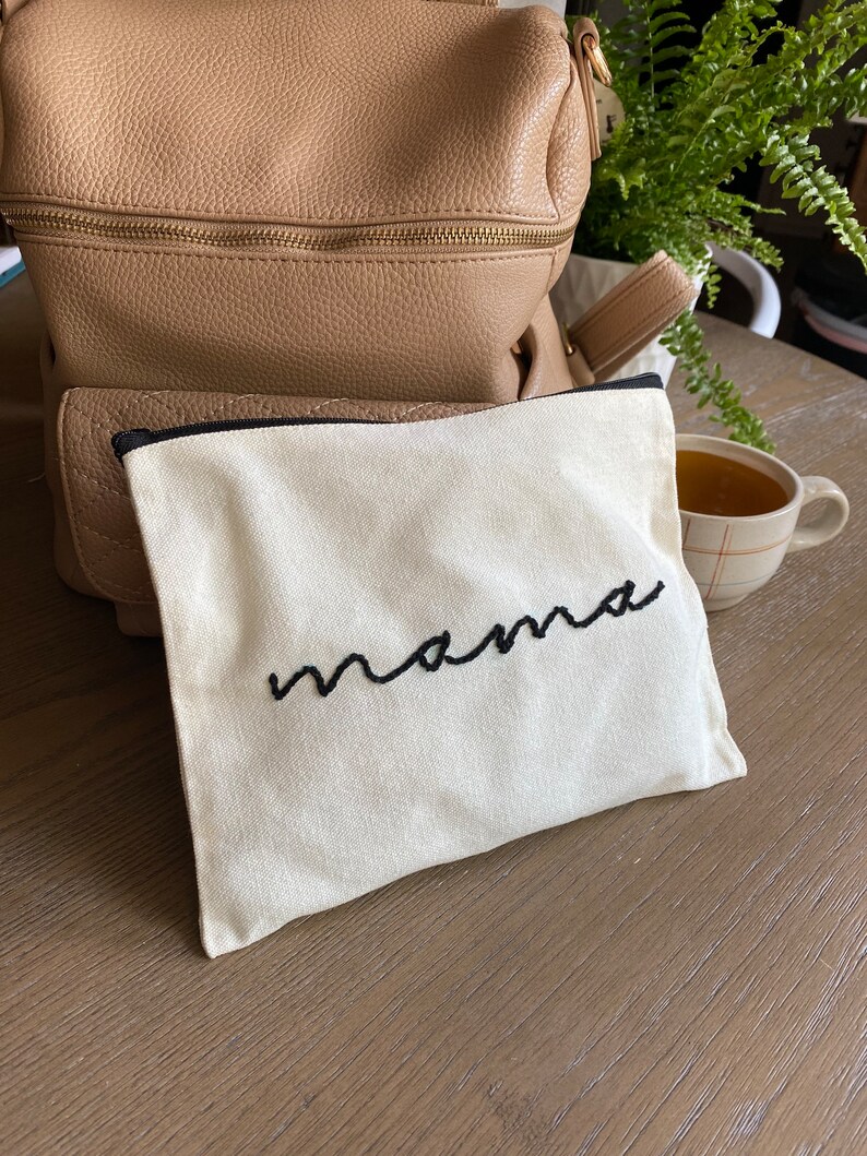 May include: A white canvas pouch with a black zipper and the word "mama" embroidered in black thread.