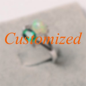 May include: A silver ring with two gemstones, one emerald green and the other iridescent. The word "Customized" is written in orange across the image. The ring is set against a blurred, neutral background.