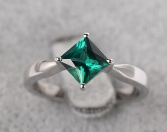May include: A silver ring featuring a square-cut, vibrant green gemstone. The gemstone is set with four prongs, and the band has a twisted design. The ring is displayed against a soft, neutral background.