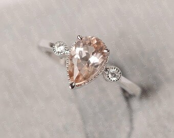 May include: A silver ring featuring a pear-shaped, peach-colored gemstone as the centerpiece. The ring is accented with smaller, round, clear gemstones on either side of the main stone, set against a light background.