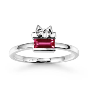 May include: A silver ring featuring a gift box design. The ring has a rectangular, deep red gemstone at the center, framed by a bow. The band is smooth and polished, with "G14K" engraved on the side. A piece of jewelry.