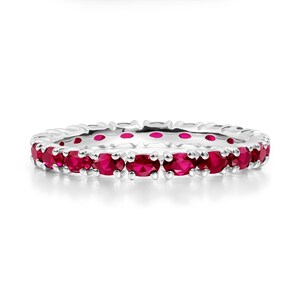 May include: A silver ring with a continuous line of oval-cut, bright red gemstones. The gemstones are set closely together, creating a full eternity band design. The ring is shown against a white background.