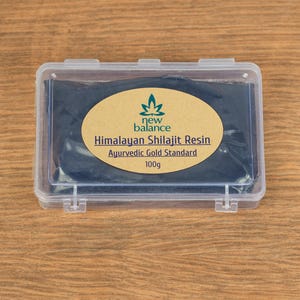 May include: A clear plastic container with a black, rectangular block inside. The block has a label that reads "new balance Himalayan Shilajit Resin Ayurvedic Gold Standard 100g".