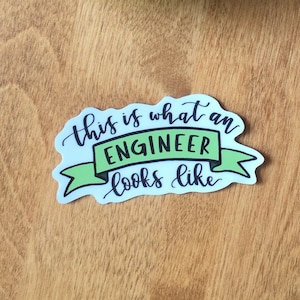 May include: A white sticker with green lettering that reads "This is what an ENGINEER looks like". The sticker is on a wooden surface with two green leaves.
