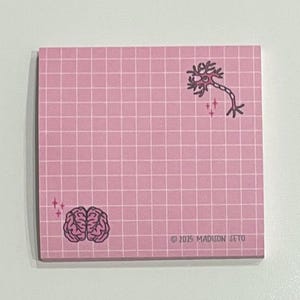 Puede incluir: Bloc de notas cuadrado rosa con un patrón de cuadrícula. El bloc de notas presenta una ilustración de cerebro y neurona, junto con el texto "© 2015 MADISON SETO". Las ilustraciones son en negro y el bloc tiene un fondo rosa claro.