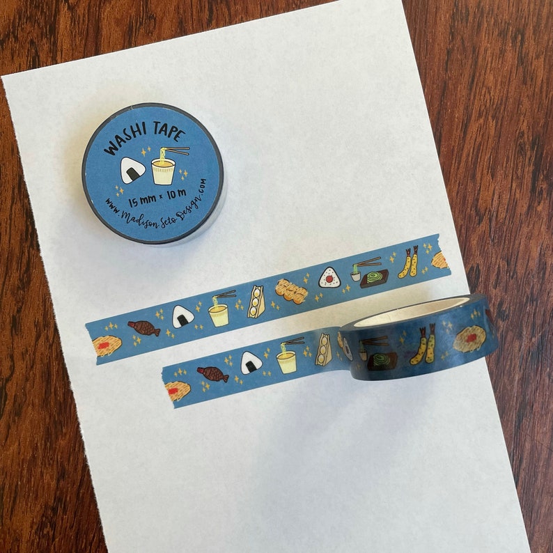 May include: A roll of blue washi tape with a repeating pattern of Japanese food, including onigiri, ramen, and tempura. The tape is 15 mm wide and 10 m long. The tape is on a white background with a small roll of the tape in the top left corner. The small roll has the text "Washi Tape" and "15 mm x 10 m" on it.
