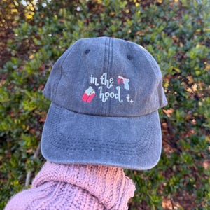May include: A charcoal grey baseball cap with a distressed finish featuring white embroidered text that reads in the hood, accompanied by a small pink juice box icon, a light blue handgun icon, and two small white cross symbols.