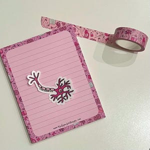 May include: A pink notepad with lined pages and a neuron sticker, alongside a roll of pink decorative tape. The tape and the notepad's border feature a repeating pattern of science-themed illustrations.
