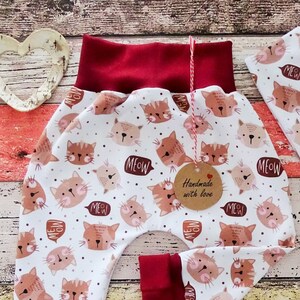 Könnte beinhalten: Rote und weiße Babyhose mit Katzenmuster und dem Wort "Meow" darauf. Die Hose hat einen roten Bund und rote Bündchen. Die Hose ist handgefertigt mit Liebe.