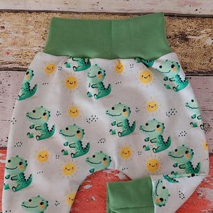 Könnte beinhalten: Babyhose mit weißem Hintergrund und einem wiederkehrenden Muster aus hellgrünen Cartoon-Krokodilen und gelben, lächelnden Sonnen. Das Kleidungsstück verfügt über einen breiten, einfarbigen salbeigrünen Elastikbund und passende Beinbündchen.