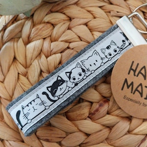 Könnte beinhalten: Ein graues Schlüsselband mit einem sich wiederholenden Muster aus schwarz-weißen Cartoon-Katzen. Das Band ist an einem silbernen Metallverschluss und einem braunen Anhänger mit der Aufschrift "HAND MADE" befestigt. Der Hintergrund ist ein gewebtes, hellbraunes Material.