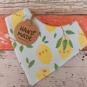 Könnte beinhalten: Ein weißes Bandana mit einem Zitronenmuster und grünen Blättern. Ein braunes Etikett mit der Aufschrift "HAND MADE" und "Especially for You" ist angebracht. Die Zitronen haben lächelnde Gesichter. Das Bandana liegt auf einer Holzoberfläche.