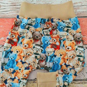 Könnte beinhalten: Eine Hose mit einem Allover-Druck aus verschiedenen Teddybären, einige davon mit Brille, in Braun-, Blau- und Weißtönen. Das Kleidungsstück hat einen breiten, einfarbigen beigen elastischen Bund und passende elastische Bündchen an den Beinabschlüssen.