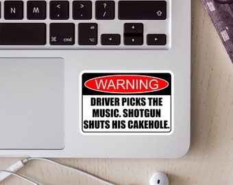 May include: A rectangular sticker with a white background and a black border. At the top, a red oval contains the white text WARNING. Below, black text reads: DRIVER PICKS THE MUSIC. SHOTGUN SHUTS HIS CAKEHOLE. The sticker is placed on a silver laptop.