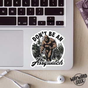May include: A sticker with a cartoon illustration of Bigfoot walking through a forest. The sticker has the text "Don't be an Assquatch" in a circle around the image.