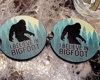 May include: Two round, blue and green coasters with a silhouette of Bigfoot walking through a forest. The coasters have the text "I believe in Bigfoot" printed on them.