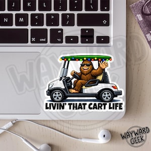 May include: A white sticker featuring a cartoon Bigfoot driving a golf cart. The Bigfoot is wearing sunglasses and waving. The golf cart has a green roof with colorful lights. The text "LIVIN' THAT CART LIFE" is below the cart.