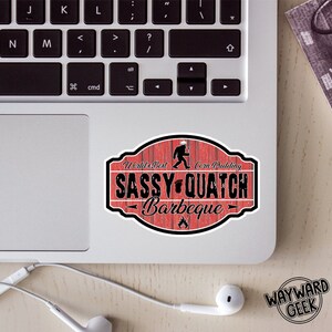 May include: An oval-shaped sticker with a red wood-plank background featuring a black silhouette of a sasquatch, a small footprint icon, and a flame graphic. The text reads World's Best Corn Pudding SASSY-QUATCH Barbeque in black lettering.
