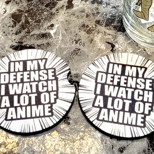 May include: Two round white coasters with black text that reads "In my defense I watch a lot of anime". The coasters have a black and white comic book style design.