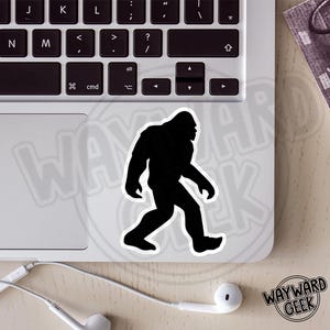 May include: A black silhouette sticker of a Bigfoot creature in a walking pose with a white border. The sticker is applied to the surface of a silver laptop keyboard area, featuring a distinct shape of the legendary cryptid in profile view.