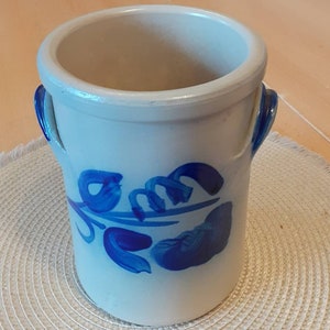 May include: A light blue ceramic vase with two small handles and a floral design in shades of blue. The vase has a cylindrical shape and sits on a woven mat.