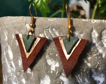 May include: A pair of handmade earrings with a geometric design. Each earring features a brown leather triangle with a green and gold accent. The earrings hang from gold-colored ear wires, showcasing a unique, artistic style.