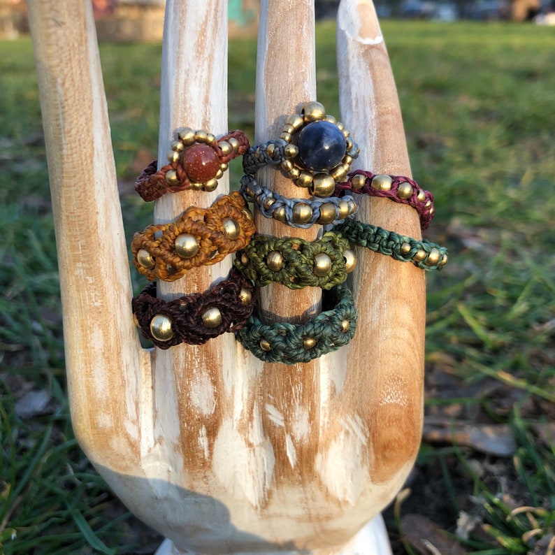 May include: A set of seven colorful macrame rings with gold beads and various stones. The rings are stacked on a wooden hand.