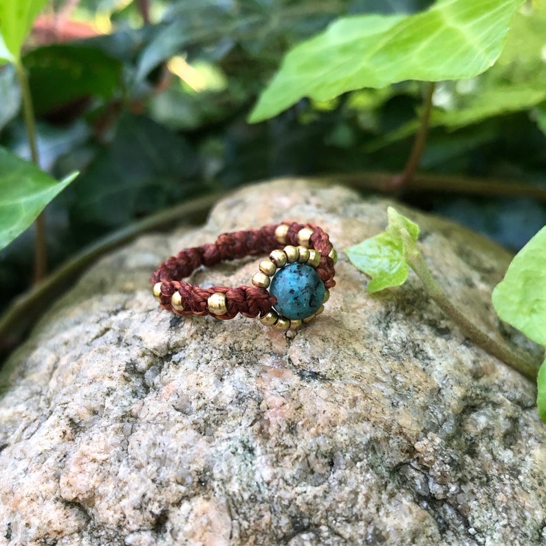 May include: A brown and gold ring with a turquoise stone. The ring is made of braided cord and has small gold beads around the stone.