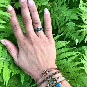 May include: A hand wearing a ring with a blue stone and several bracelets with brown cord, beads, and a metal charm with a blue stone.