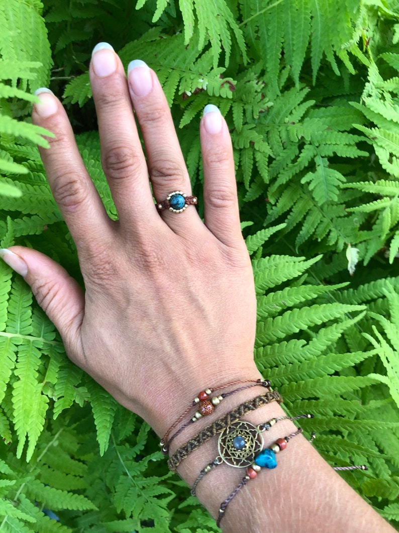 May include: A hand wearing a ring with a blue stone and several bracelets with brown cord, beads, and a metal charm with a blue stone.