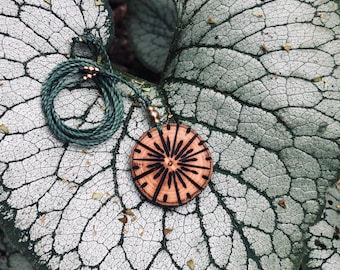 May include: A wooden pendant necklace with a sunburst design. The pendant is round, with a dark brown sunburst pattern etched into the light brown wood. The necklace has a green cord. The pendant rests on a large, textured leaf.