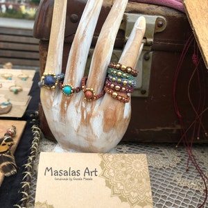 May include: A wooden hand model displaying a variety of colorful beaded rings with gemstone accents. The rings are made with natural materials and have a bohemian aesthetic. The hand model is holding a business card that reads "Masalas Art handmade by Gesala Masala" with contact information for Etsy, Instagram, and email.