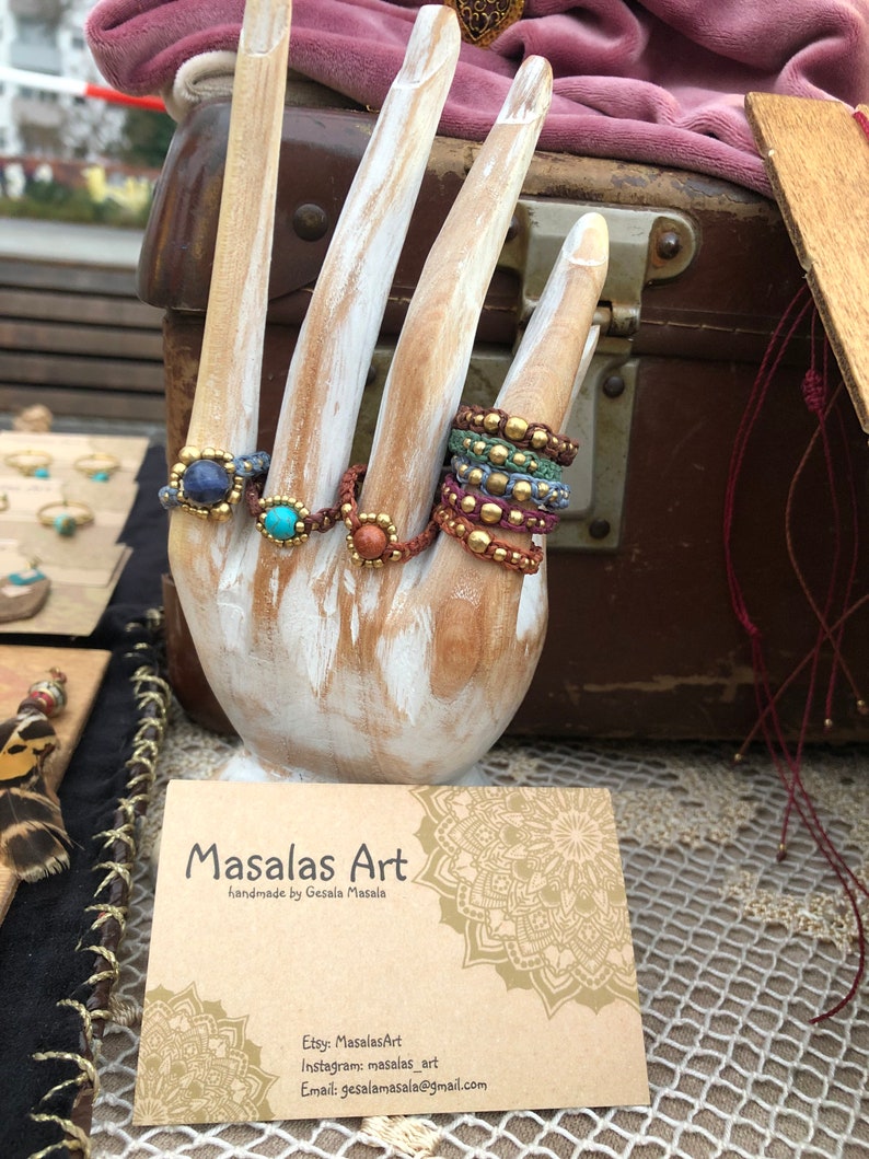 May include: A wooden hand model displaying a variety of colorful beaded rings with gemstone accents. The rings are made with natural materials and have a bohemian aesthetic. The hand model is holding a business card that reads "Masalas Art handmade by Gesala Masala" with contact information for Etsy, Instagram, and email.
