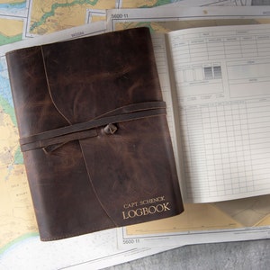 May include: A brown leather logbook with the text "Capt. Schenck Logbook" in gold lettering. The logbook is open, revealing lined pages for recording data. A leather strap wraps around the closed book. Nautical charts are in the background.