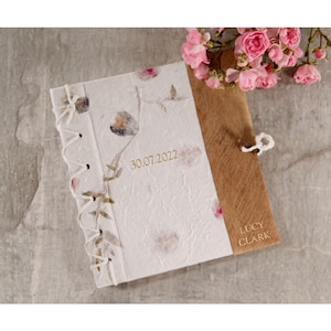 Puede incluir: Un diario blanco hecho a mano con una cubierta de madera y un cierre de encaje. El diario tiene un diseño floral y la fecha "30.07.2022" y el nombre "Lucy Clark" están grabados en la cubierta.